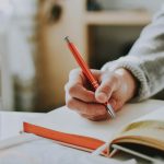 A person writing in a notebook with a red ballpoint pen, showcasing detail and focus.