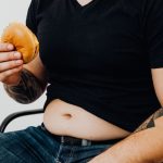 Close-up of a man holding a burger, showcasing his torso and tattooed arm. Casual and relaxed setting.