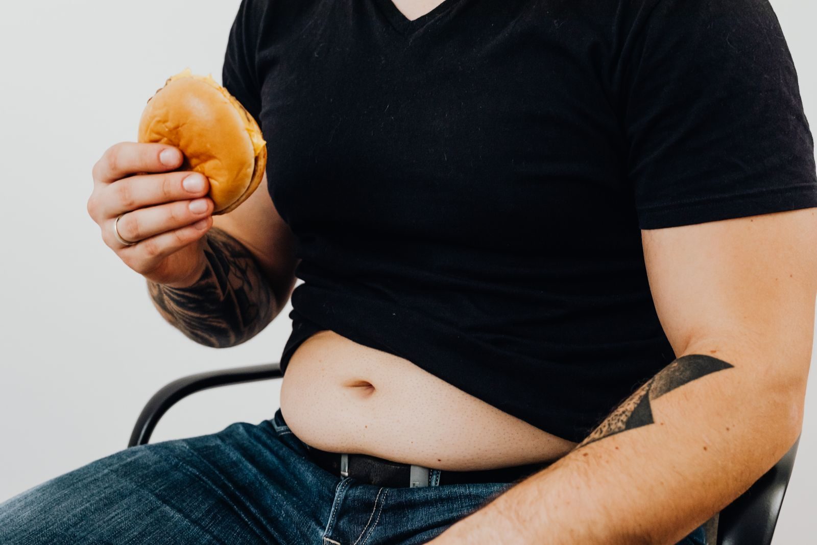 Close-up of a man holding a burger, showcasing his torso and tattooed arm. Casual and relaxed setting.